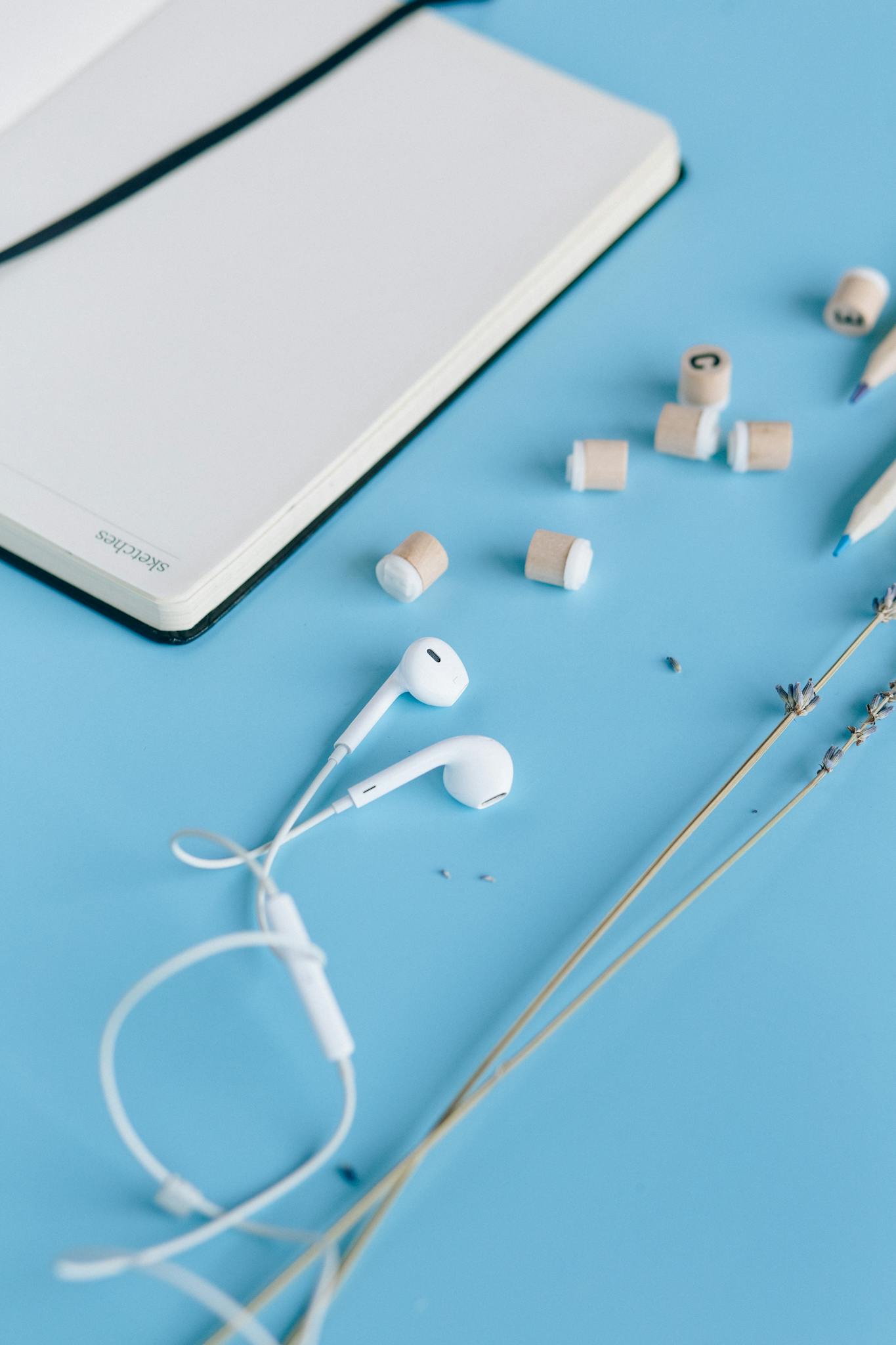 Flat lay of a minimalist workspace with a notebook, earbuds, and writing tools on a blue background.